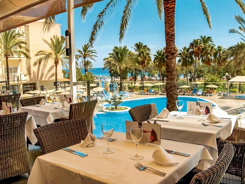 Buitenrestaurant met zitplaatsen bij zwembad met palmbomen en uitzicht op zee in zonnig resort