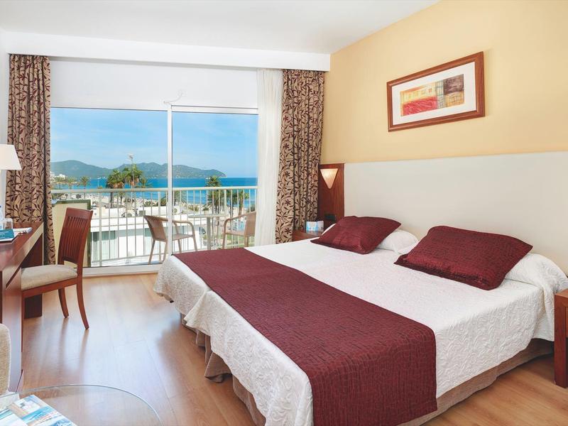 Lichte hotelkamer met tweepersoonsbed, balkon en uitzicht op zee door grote glazen deuren.