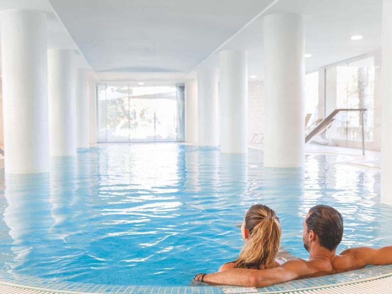 Koppel ontspant in een ronde overdekte zwembad met witte pilaren en ramen.