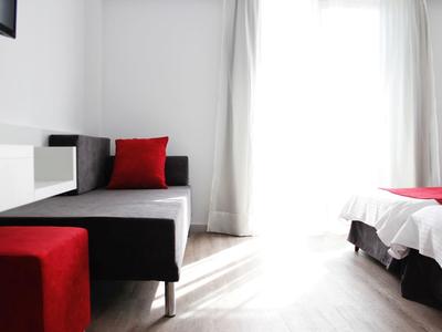 Helles Schlafzimmer mit weißem Bett, rotem Hocker und rotem Kissen, großer Fensterlichtquelle.