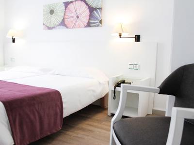 Hell eingerichtetes Schlafzimmer mit Doppelbett, weißen Wänden, Holzboden und grauen Sitzmöbeln.