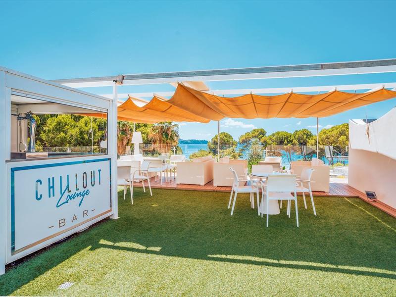 Außenbereich einer Strandbar mit weißen Tischen unter orangefarbenem Sonnenschutz und Blick auf das Meer.