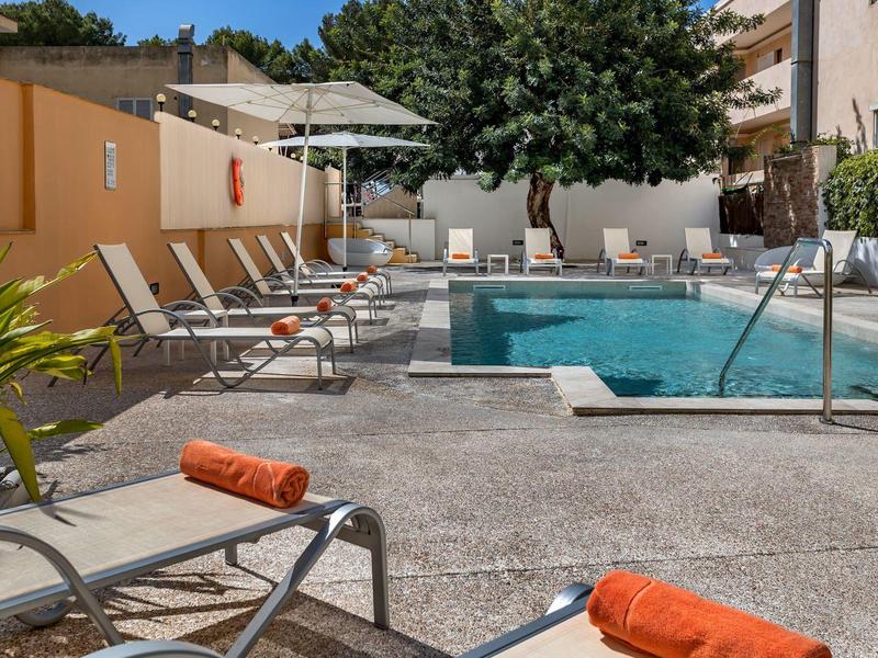 Hotelpool mit Liegestühlen, Sonnenschirmen und Baum im Hintergrund an einem sonnigen Tag.