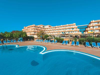 Large outdoor pool with blue lounge chairs and a hotel building in the background under clear sky.