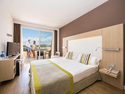 Helles Hotelzimmer mit Doppelbett, TV, Sitzbereich und Balkon mit Bergblick.