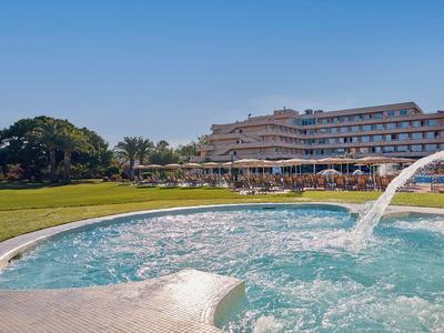 Großes Hotel mit Pool, Wasserfontäne und Sonnenliegen auf grünem Rasen unter blauem Himmel.