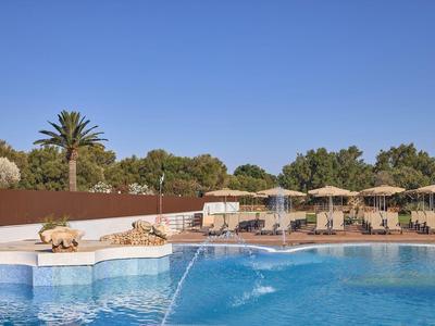 Großer Pool mit Wasserspielen und Liegestühlen unter Sonnenschirmen in einer Urlaubsanlage.