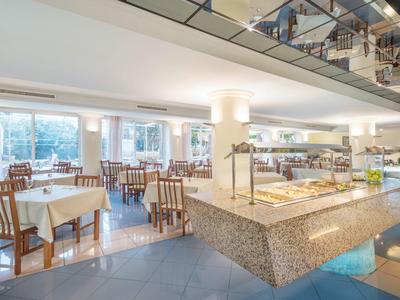 Světlá hotelová restaurace s bufetovou částí a několika stoly se židlemi.