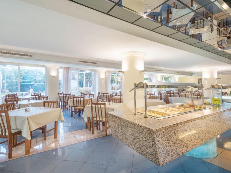 Světlá hotelová restaurace s bufetovou částí a několika stoly se židlemi.