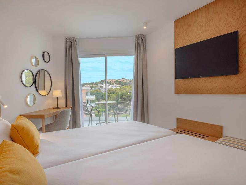 Moderní hotelový pokoj se dvěma postelemi, žlutými polštáři, zrcadlovou zdí a balkonem s výhledem.