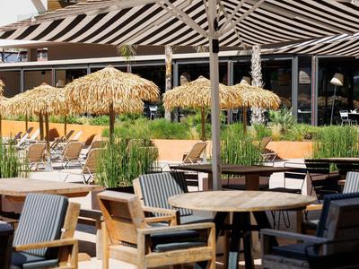 Area esterna di un hotel con tavoli e sedie in legno e ombrelloni di paglia sullo sfondo.