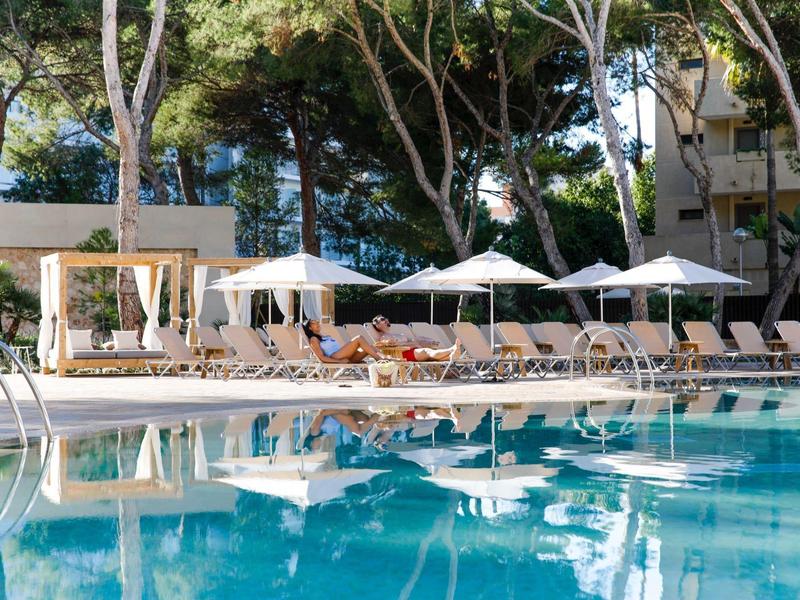 Area piscina con lettini, ombrelloni e cabana in un giardino di un hotel.