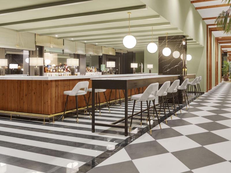 Area bar dell'hotel moderna con bancone in legno, sgabelli bianchi e pavimento a scacchi bianco e nero.