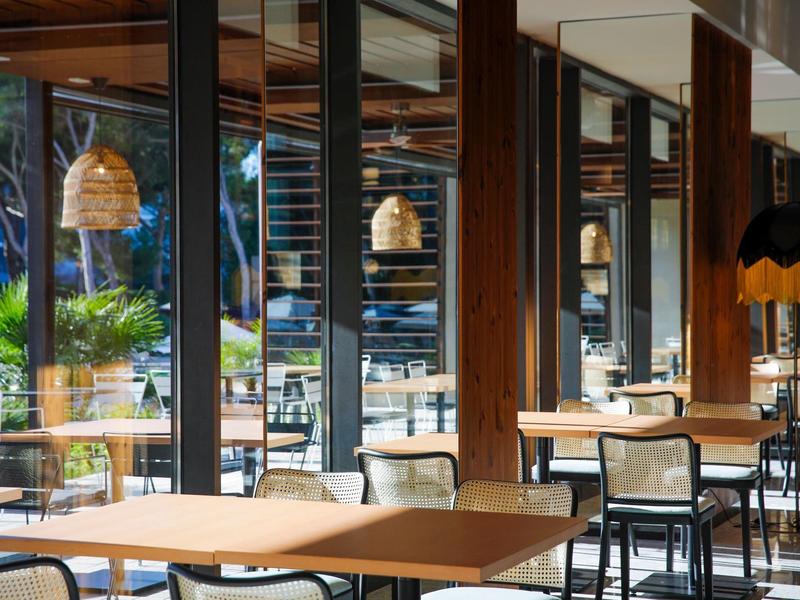 Interno di un ristorante moderno e luminoso con mobili in legno e grandi finestre in vetro.