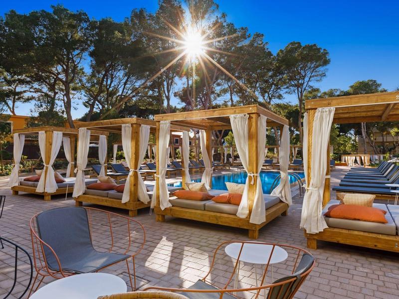 Lussuosa lounge esterna con lettini e tende da sole sotto gli alberi in una giornata di cielo sereno.