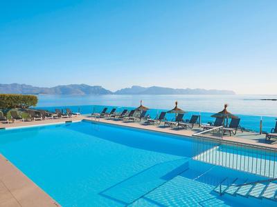 Großer, klarblauer Pool mit Liegestühlen und Blick auf das Meer und Berge im Hintergrund bei Sonnenschein.
