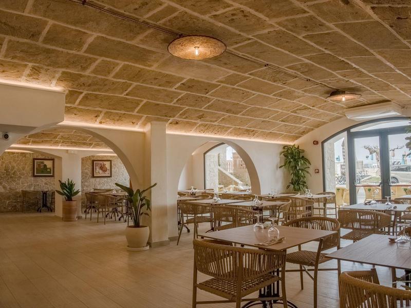Ristorante d'hotel luminoso con archi, sedie e tavoli in legno, grandi finestre e piante.