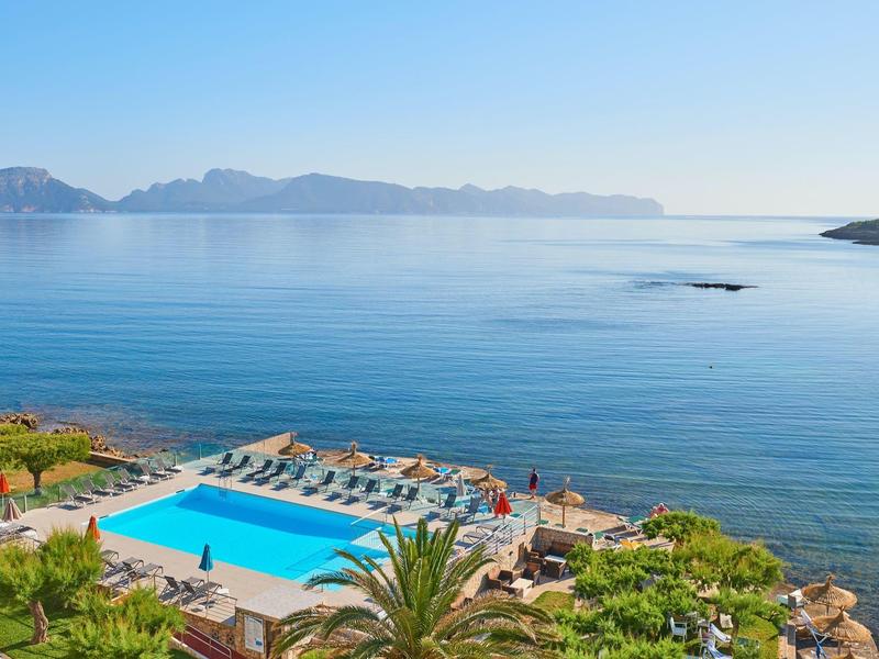 Piscina con lettini a bordo mare con vista su una baia tranquilla e montagne sullo sfondo.