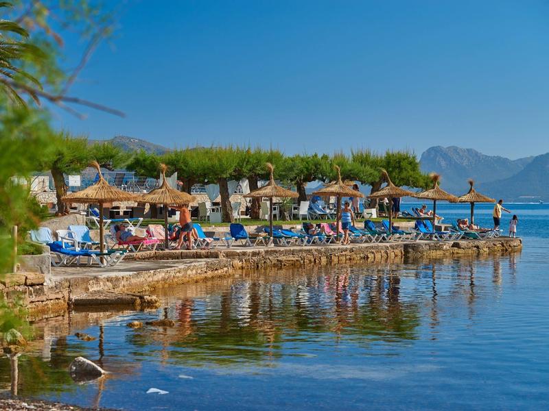 Passeggiata sulla spiaggia con lettini, ombrelloni e montagne sullo sfondo vicino ad acqua calma.