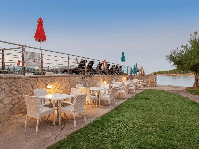 Area terrazza con tavoli, sedie e lettini accanto a prato verde e vista sull'acqua.