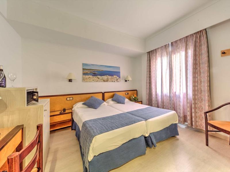 Bright hotel room with two single beds, nightstands, and a window with curtains.