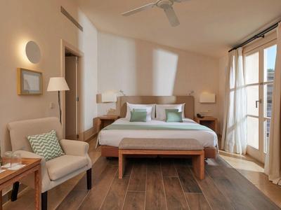 Pohodlný hotelový pokoj s manželskou postelí, křeslem, dřevěnou podlahou a balkónovými dveřmi se závěsy.