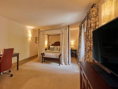 Pohodlný hotelový pokoj s postelí s nebesy, psacím stolem, televizí a teplým osvětlením.