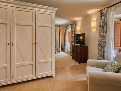 Prostorný hotelový pokoj s velkou skříní, křeslem, komodou, TV a oknem se záclonami.