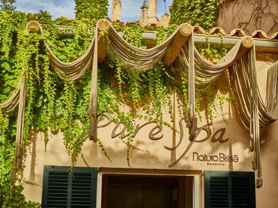 Vstup do lázní zdobený zavěšenými lany a bujnými zelenými rostlinami za slunečného dne.