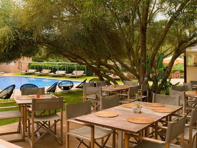 Venkovní jídelní prostor s dřevěnými stoly a židlemi u bazénu obklopeného zelení.