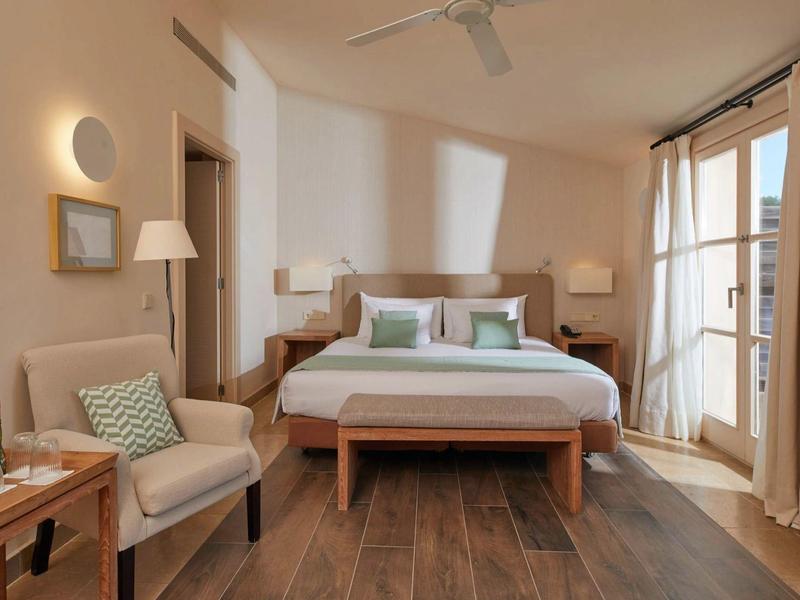 Pohodlný hotelový pokoj s manželskou postelí, křeslem, dřevěnou podlahou a balkónovými dveřmi se závěsy.