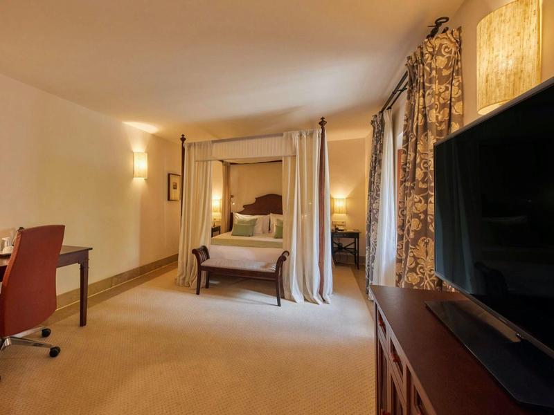 Pohodlný hotelový pokoj s postelí s nebesy, psacím stolem, televizí a teplým osvětlením.