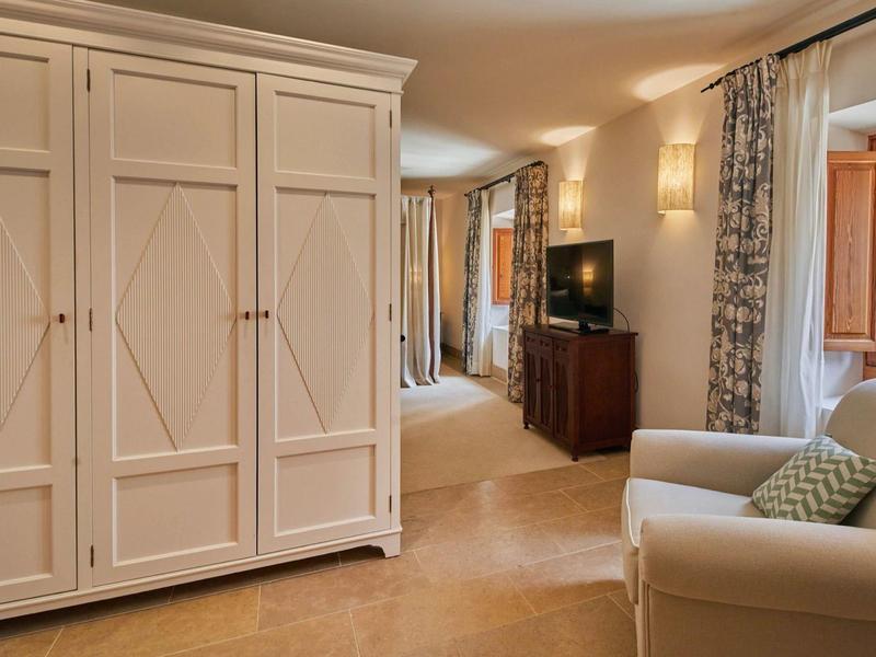 Prostorný hotelový pokoj s velkou skříní, křeslem, komodou, TV a oknem se záclonami.