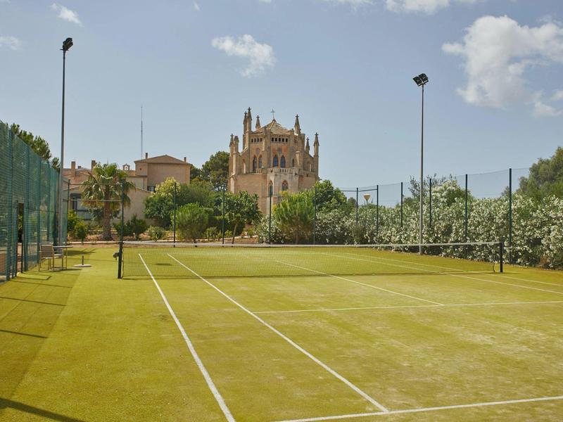 Travnatý tenisový kurt obklopený oplocením s historickou budovou v pozadí pod modrou oblohou.