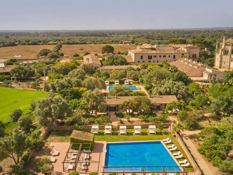 Letovisko s velkým modrým bazénem, lehátky, zelenými stromy, poli a budovami pod jasnou oblohou.