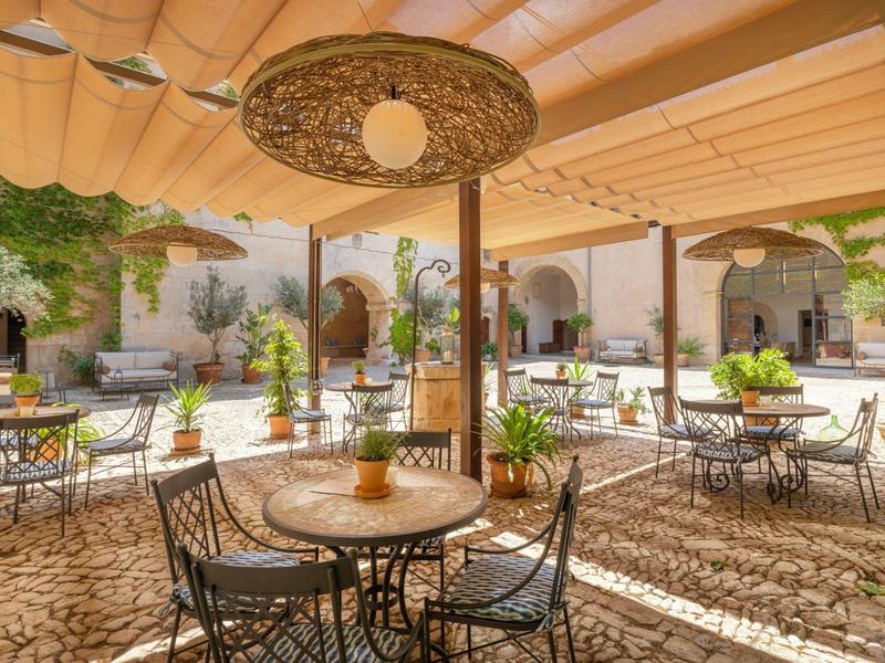 Zastíněné venkovní nádvoří s kulatými stoly, železnými židlemi a kamennou podlahou.