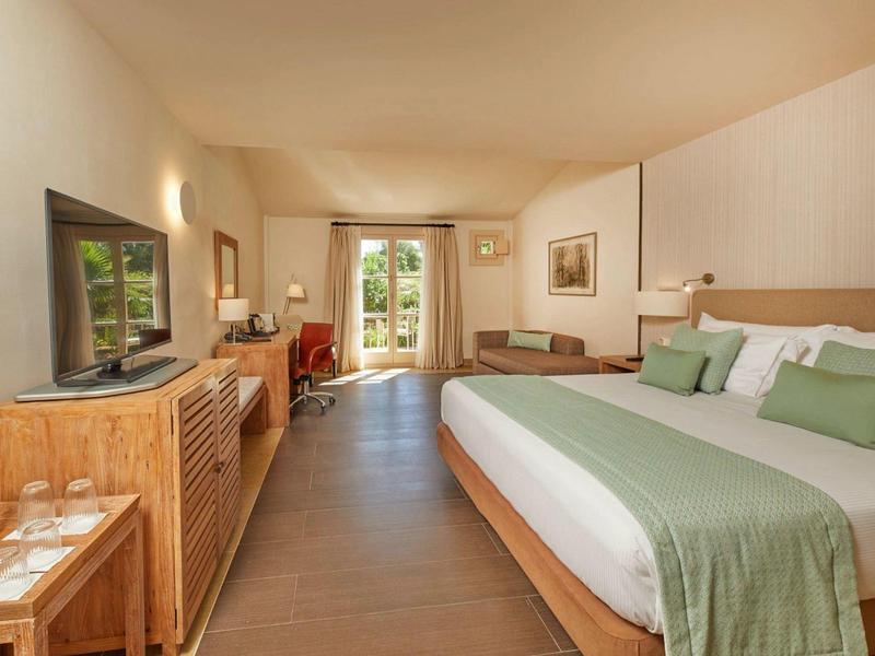 Prostorný hotelový pokoj s velkou postelí, dřevěným nábytkem a výhledem do zahrady.