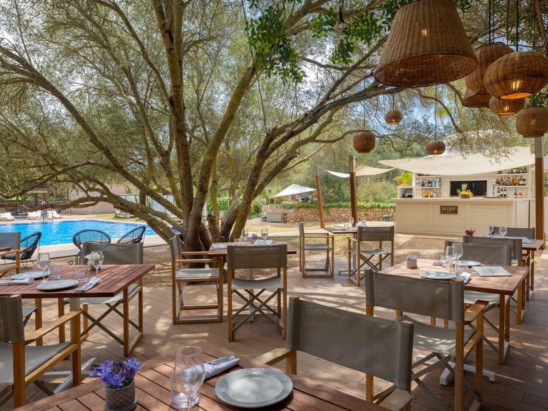 Venkovní restaurace s dřevěnými stoly a bazénem pod velkými stromy.