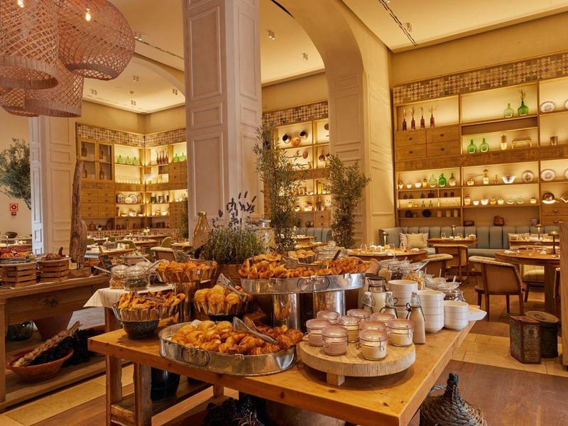 Příjemná pekárenská část s dřevěnými stoly plnými chleba, pečiva a sklenic v světlé kavárně.