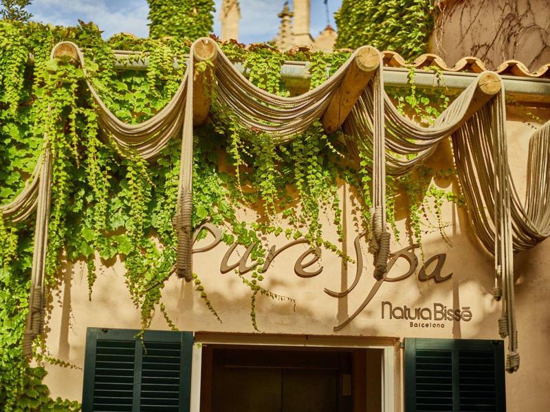 Vstup do lázní zdobený zavěšenými lany a bujnými zelenými rostlinami za slunečného dne.