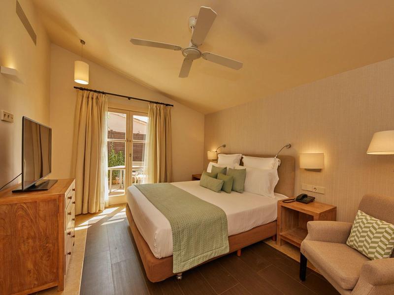 Pohodlný hotelový pokoj s manželskou postelí, dřevěným nábytkem, křeslem a balkonem.