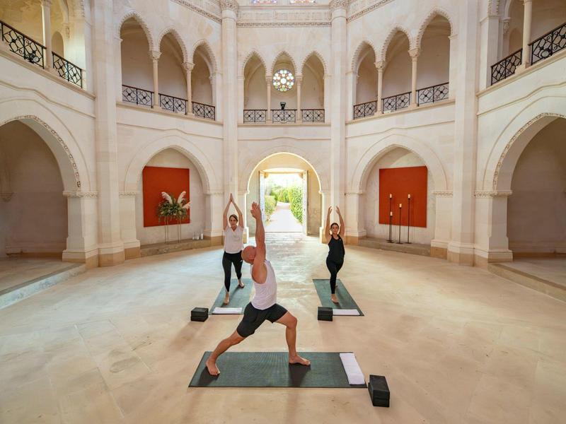 Tři lidé cvičí jógu ve velkém elegantním vnitřním prostoru s oblouky a světlými stěnami.