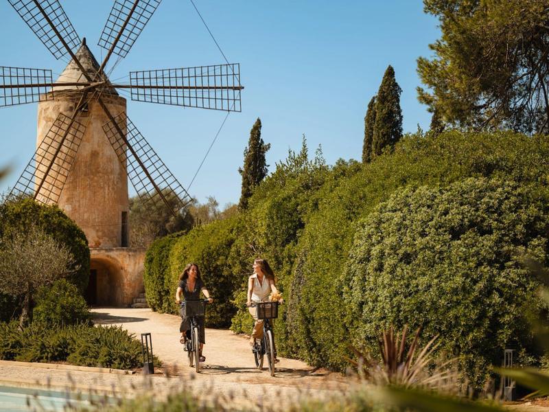 Dva lidé jedou na kole stezkou vedle zelených keřů a starého větrného mlýna pod jasnou oblohou.
