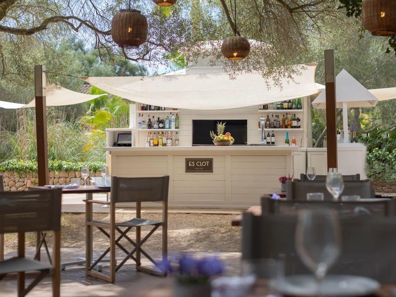 Venkovní bar s bílým přístřeškem, dřevěnými židlemi a zavěšenými lucernami obklopený stromy.