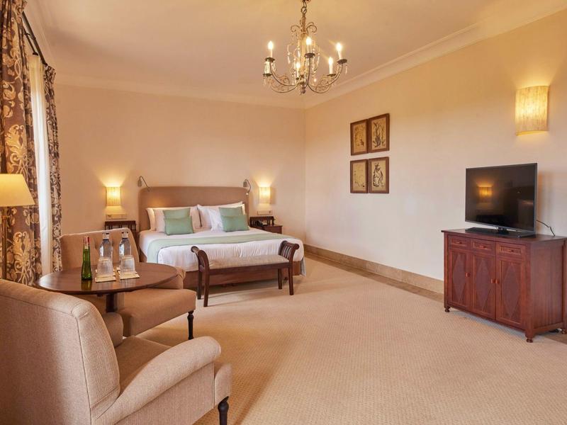 Pohodlný hotelový pokoj s postelí, křeslem, dřevěnou skříní, televizí a teplým osvětlením.