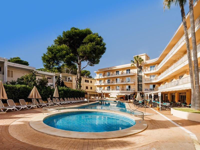 Un hotel con una piscina circular y tumbonas bajo un cielo despejado y soleado.