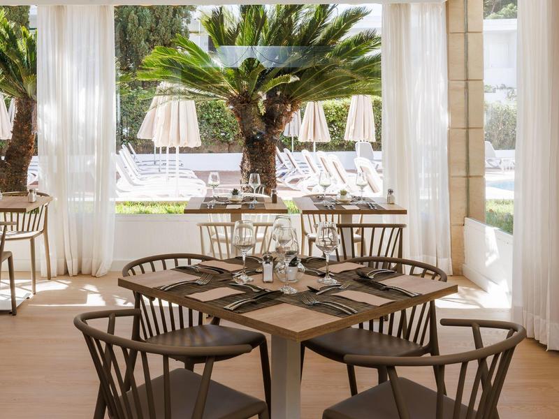 Área exterior elegante de un hotel con mesas de madera, sillas y fondo de plantas tropicales.