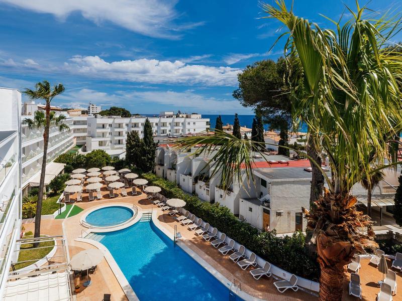 Piscina de hotel con hamacas y palmeras bajo un cielo azul junto al mar.