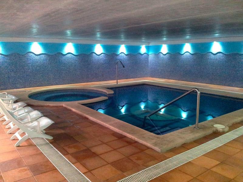Piscina interior con agua azul, suelo de terracota, tumbonas blancas y ambiente tranquilo en un hotel.