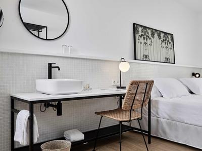 Moderní hotelový pokoj s bílou postelí, dřevěnou židlí a nástěnným umyvadlem.